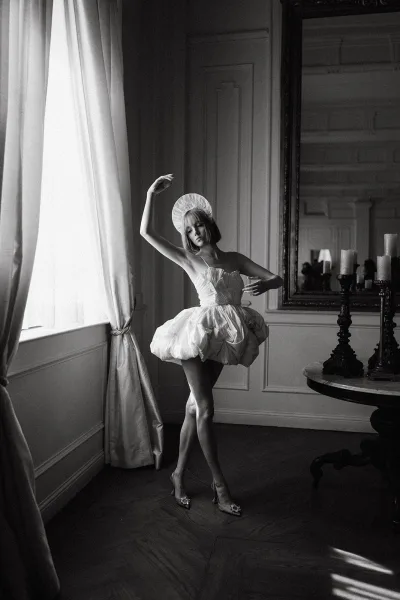 Bridal portrait of a bride in a short wedding dress with structured headpiece and heels, posed by a mirror in window light and candles