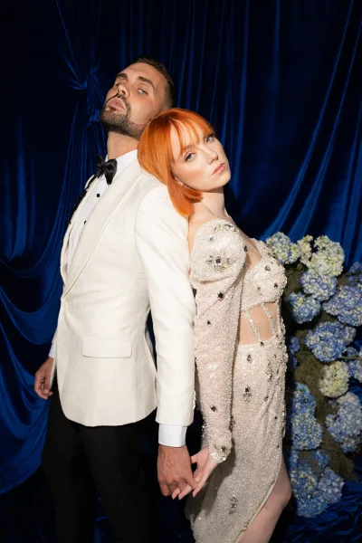 Couple portrait of bride in a beaded wedding dress leaning on groom in a white dinner jacket, holding hands before a blue velvet drape backdrop