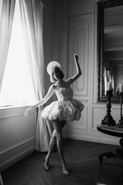 Bridal portrait of a bride in a short wedding dress and headpiece by window light, reflected in a wall mirror amid paneled walls and candles