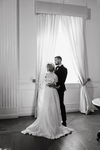 Couple portrait, black and white wedding portrait of groom hugging bride in strapless dress and headpiece by large window with curtains