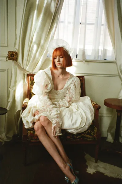 Bridal portrait of a redhead bride seated on a vintage armchair in a lace-sleeve gown, wearing blue heels by a window with white curtains