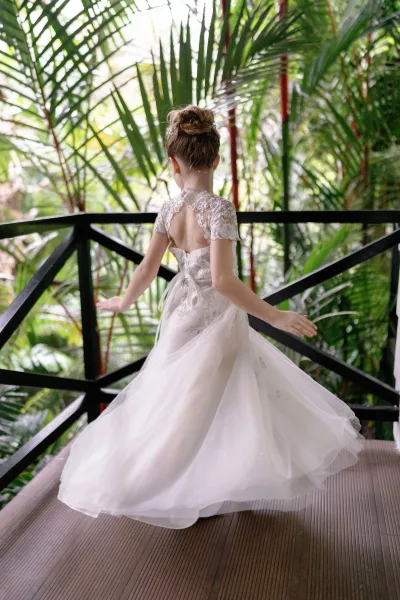Flower girl portrait of a twirling flower girl in an ivory dress with a lace bodice on a wooden deck framed by tropical plants.