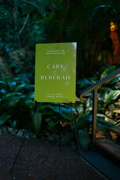 Wedding welcome sign, a green wedding sign on an easel stand, framed by tropical foliage along a garden path beside a staircase railing.