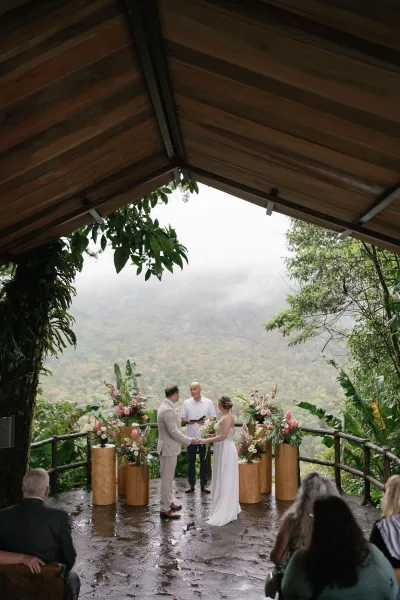 Ceremony moment as the couple exchange outdoor wedding vows, framed by wooden pedestal florals, with rainforest foliage and a misty mountain view.