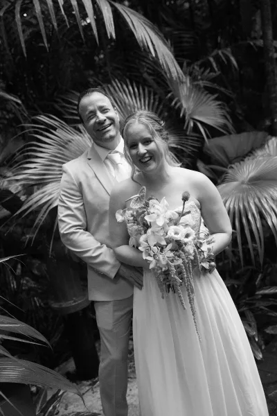 Couple portrait of bride and groom smiling, her orchid bridal bouquet between them, set against lush palm fronds in a tropical garden.