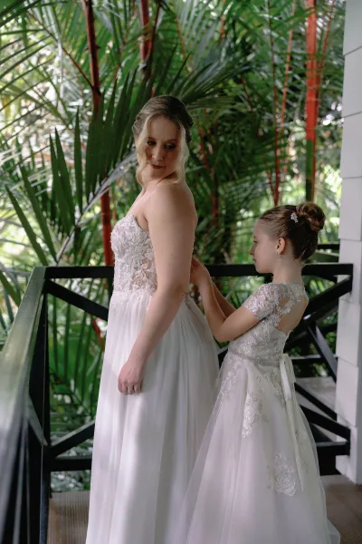 Bride getting ready with flower girl buttoning her sheer lace appliqué bodice on a porch, framed by tropical greenery and white siding.