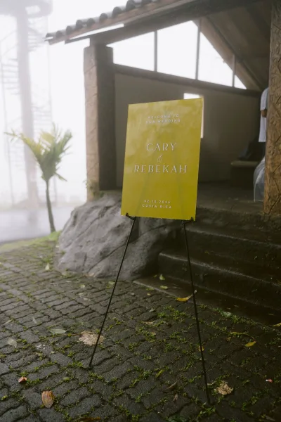 Wedding welcome sign in modern wedding signage style on an easel stand along a cobblestone path, framed by stone steps and a foggy outdoor scene.
