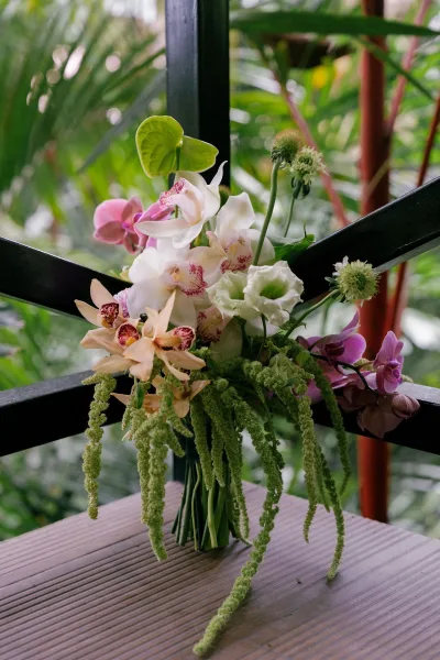 Bridal bouquet in an orchid wedding bouquet style, accented with trailing amaranthus, resting on a wood tabletop before tropical foliage.