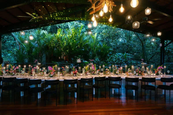 Reception tablescape on a long banquet table with glowing candles, set under a pavilion amid tropical plants and palm trees.