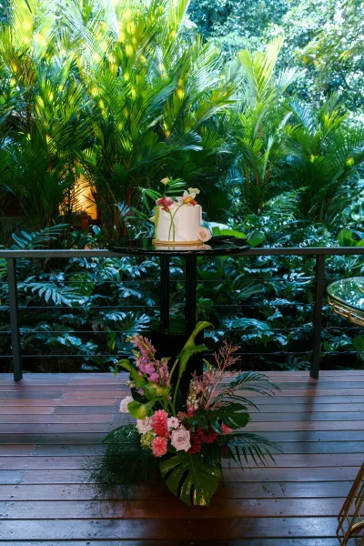 Wedding cake, single tier wedding cake in smooth buttercream topped with a pink rose on a wooden deck amid lush tropical garden greenery.