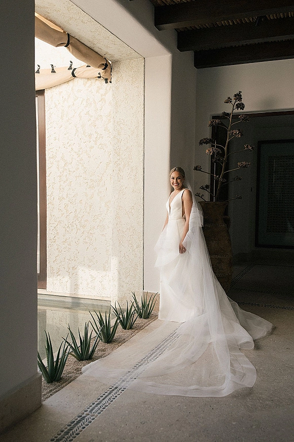 Bridal portrait of a bride in a simple wedding dress with a long veil and train, standing by a stucco patio wall with desert plants