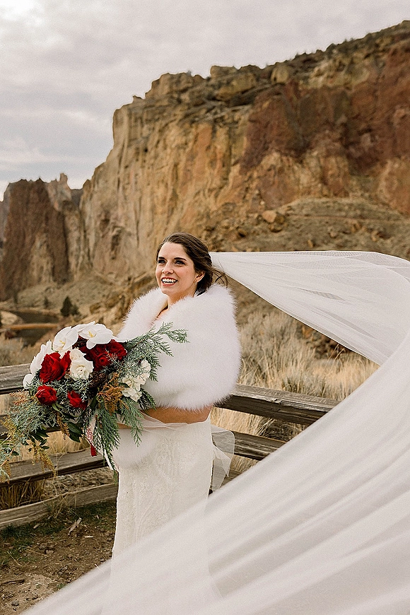 Bridal portrait of a bride holding bouquet, veil blowing in the wind as she stands by rock cliffs and a wooden fence under gray skies