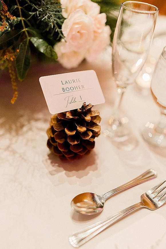 Wedding place card with calligraphy nestled in a pine cone holder beside a champagne flute on a white linen reception tablescape with greenery