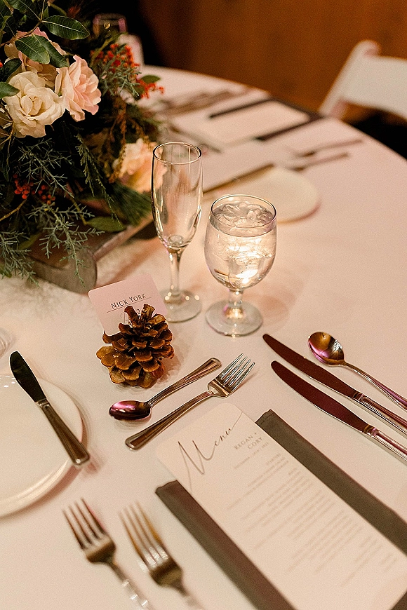 Reception tablescape with wedding place setting, floral centerpiece and greenery, menu cards and pine cone place card holder on runner by wooden wall