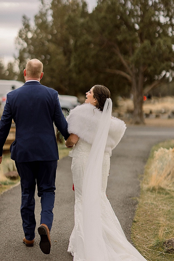 Couple portrait of bride and groom walking away on a tree-lined road, her long veil and lace dress with a fur wrap, his navy suit