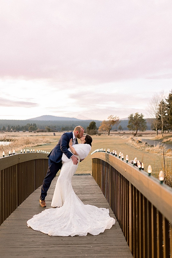 Wedding kiss portrait of newlyweds in a dip on a wooden bridge, bride in fur wrap and long train, mountains and cloudy sky behind