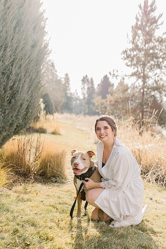 Bridal portrait of a bride in a lace robe kneeling with her dog on a leash in tall grass, sunlight and evergreen trees behind them