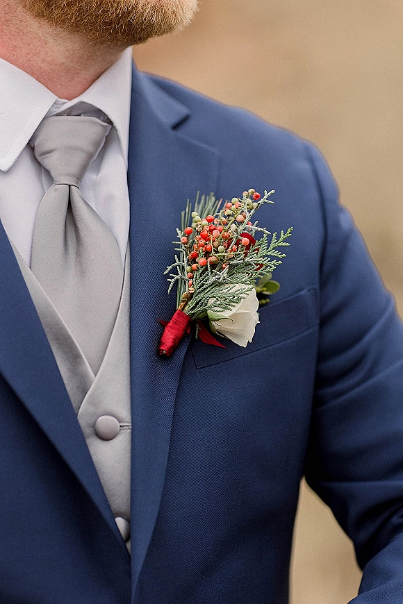Groom boutonniere with a white rose and red berries nestled in evergreen foliage, wrapped in red ribbon on a blue suit jacket