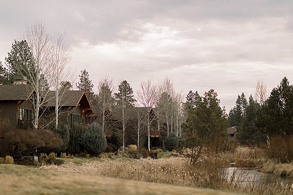 Rustic lodge exterior with wood cabin buildings and pitched roofs, featuring a covered porch and chimney beside a creek under cloudy sky