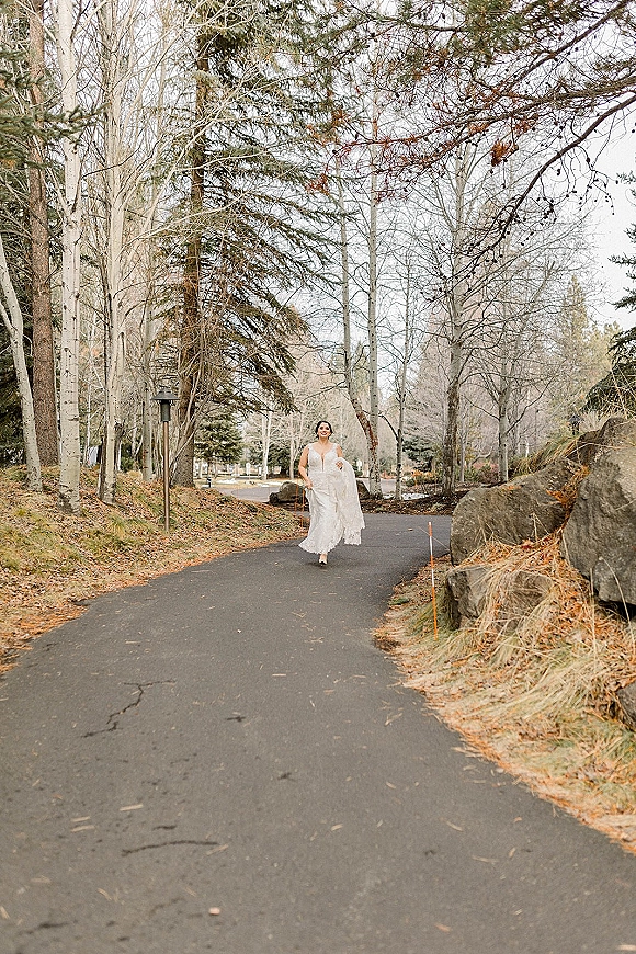 Bridal portrait of a bride in a lace gown holding up her tulle skirt while running along a forest path under an overcast sky