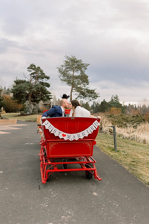 Wedding kiss portrait of newlyweds in carriage, a red horse-drawn getaway with a just married banner on a country road under clouds