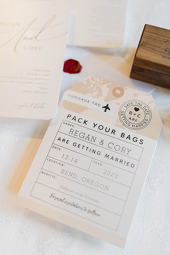 Save the date card with travel themed save the date details, luggage tag and passport stamp graphics on white linen, red rose petal nearby