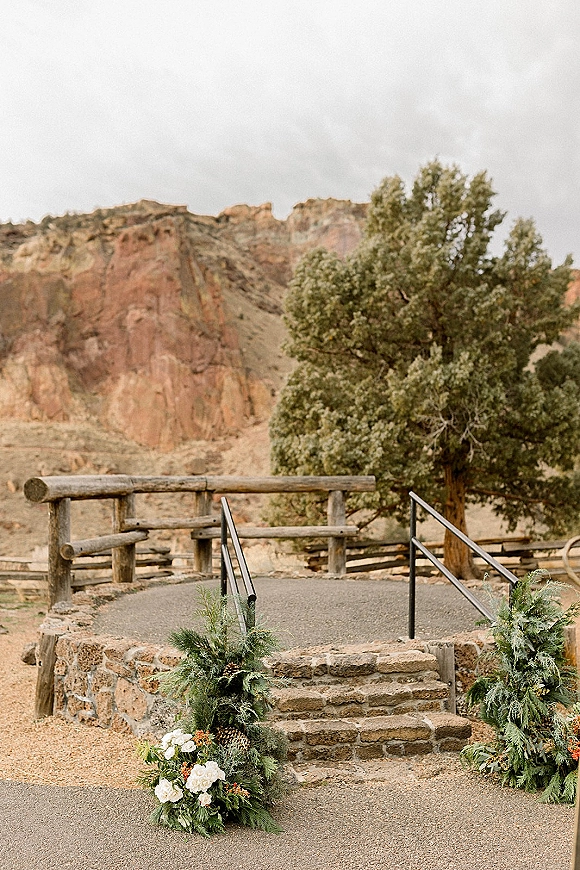 Ceremony entrance decor with evergreen garland and white flowers, pinecones and orange blooms lining stone steps under desert cliffs