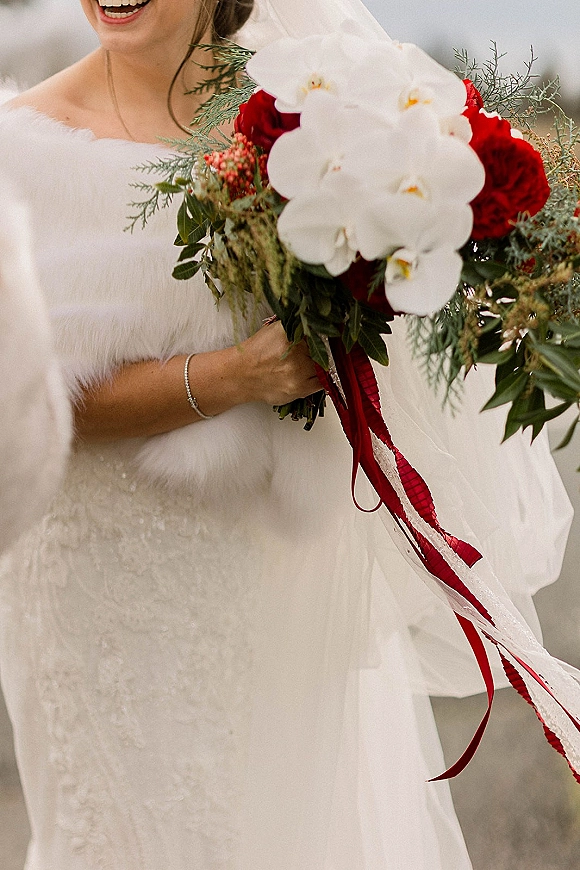 Bridal bouquet of white orchids and red roses with greenery and ribbon, held by a bride in a beaded strapless gown and veil, by a neutral wall
