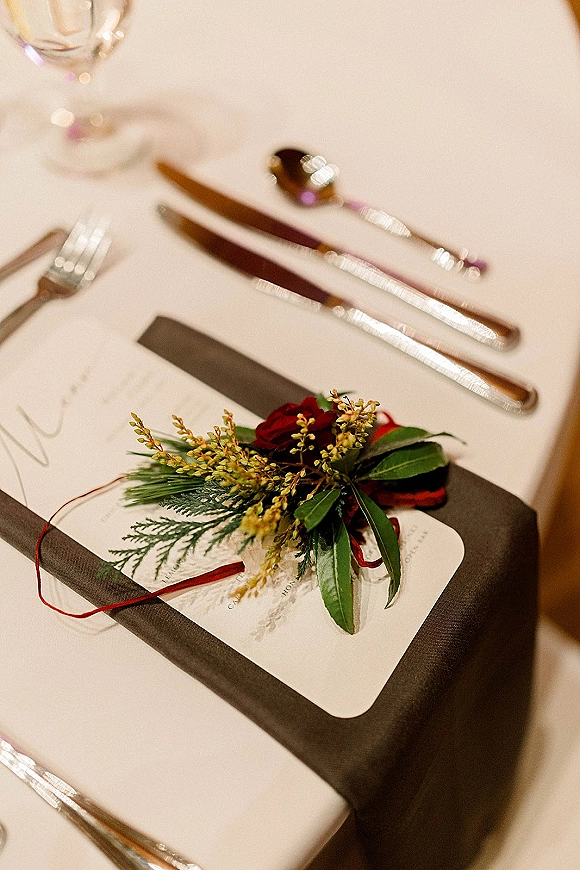 Wedding place setting with wedding menu card on a folded napkin, red ribbon wrap, gold flatware and glassware on a white tablecloth