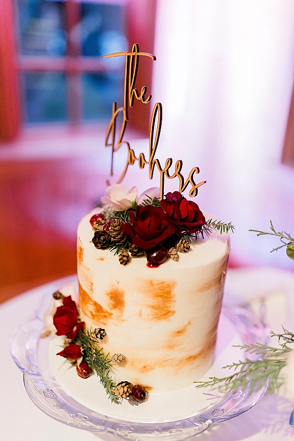 Wedding cake with semi naked frosting topped with gold script topper and red roses, evergreen sprigs and pinecones by a window indoors