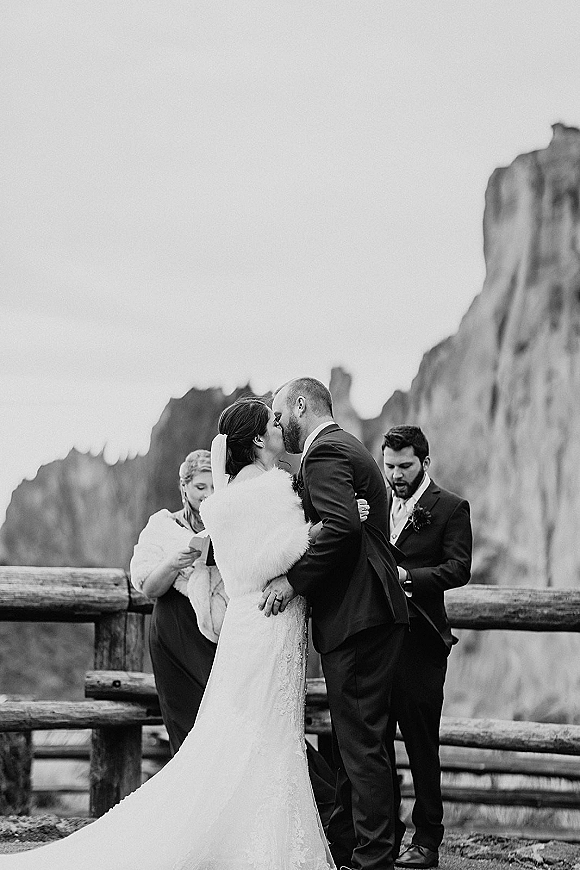 Wedding kiss as the couple embraces in a black and white ceremony moment, bride in veil and faux fur shawl by mountain cliffs