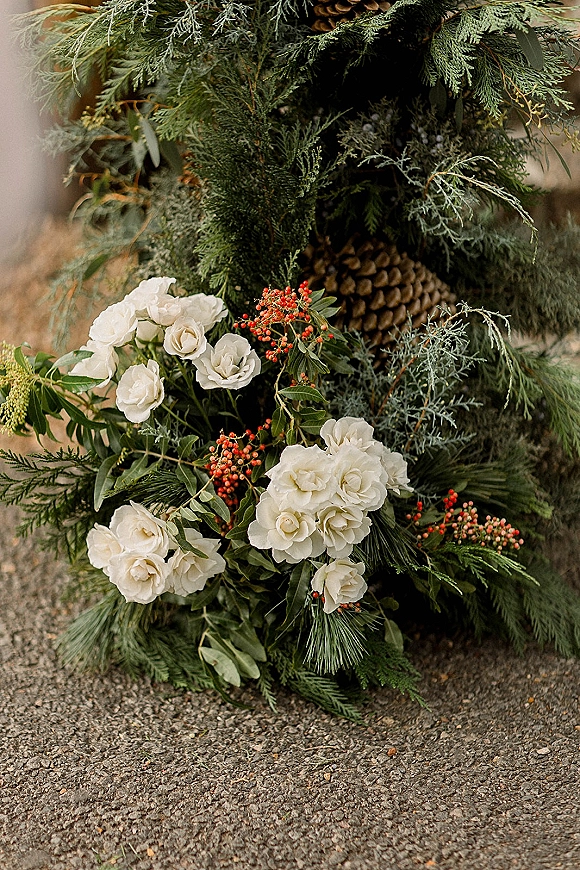 Wedding ceremony decor with evergreen ceremony garland of white roses, red berries, pinecones, and greenery on asphalt outdoors
