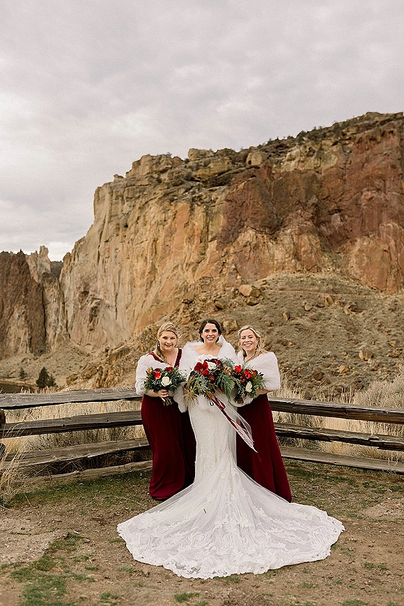 Bride with bridesmaids portrait shows her in a lace train and faux fur wrap, bridesmaids in burgundy holding red and white bouquets by canyon cliffs