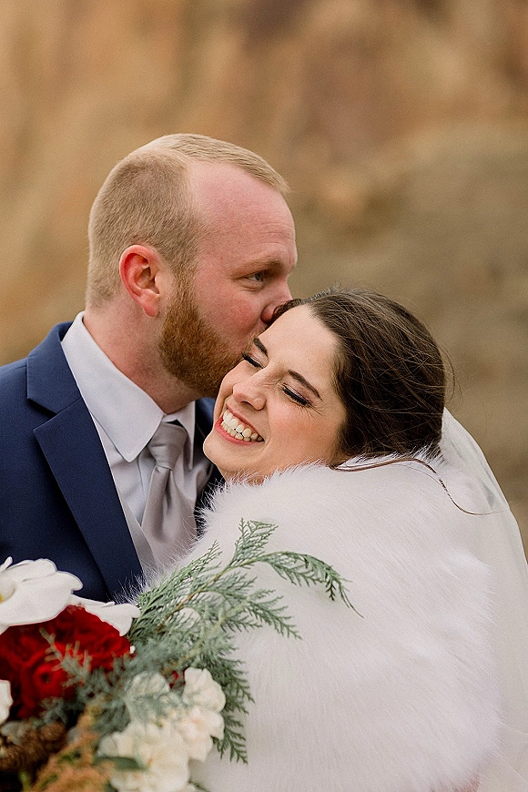 Wedding couple portrait with groom kissing bride’s forehead as she smiles, holding a red rose bouquet and wearing a faux fur wrap among blurred trees