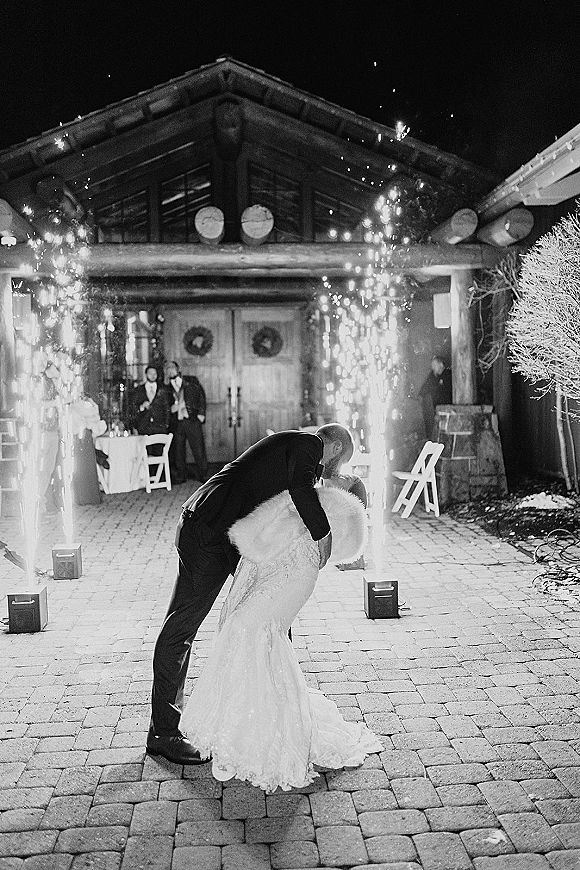 Wedding kiss portrait of bride in lace dress and faux fur wrap dipped by tuxedo groom as sparkler fountains light rustic lodge entrance at night