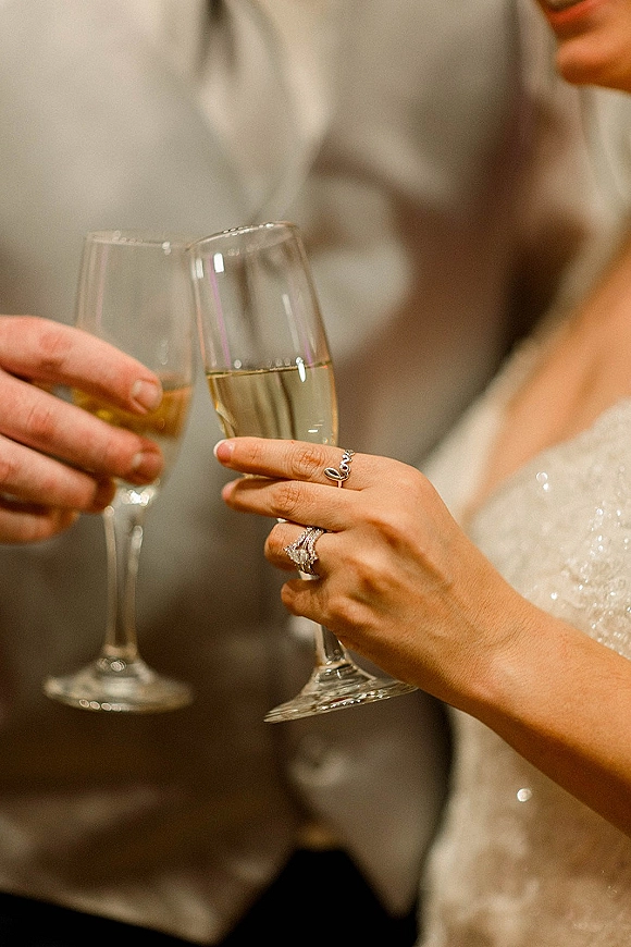 Wedding toast with bride and groom clinking champagne flutes, rings visible on hands during an indoor reception against a neutral wall