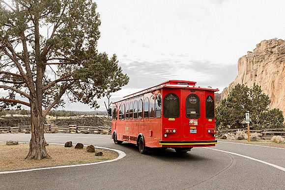 Red trolley bus wedding shuttle bus driving along a curved desert canyon road, with pine trees, wooden fence, and rock cliffs under overcast sky