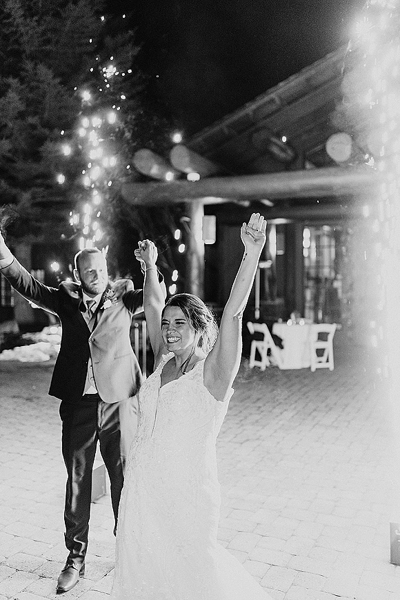 Wedding sparkler exit with bride raising her arms as groom cheers behind her, sparklers glowing under string lights on an outdoor patio at night