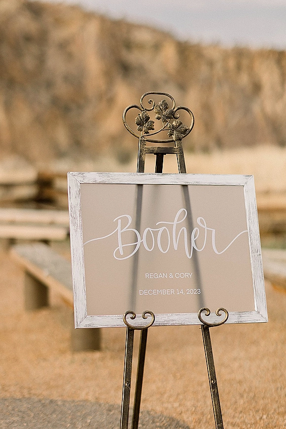 Wedding welcome sign with calligraphy lettering in a framed board on a metal easel, set against mountains, fence, and gravel ground outdoors