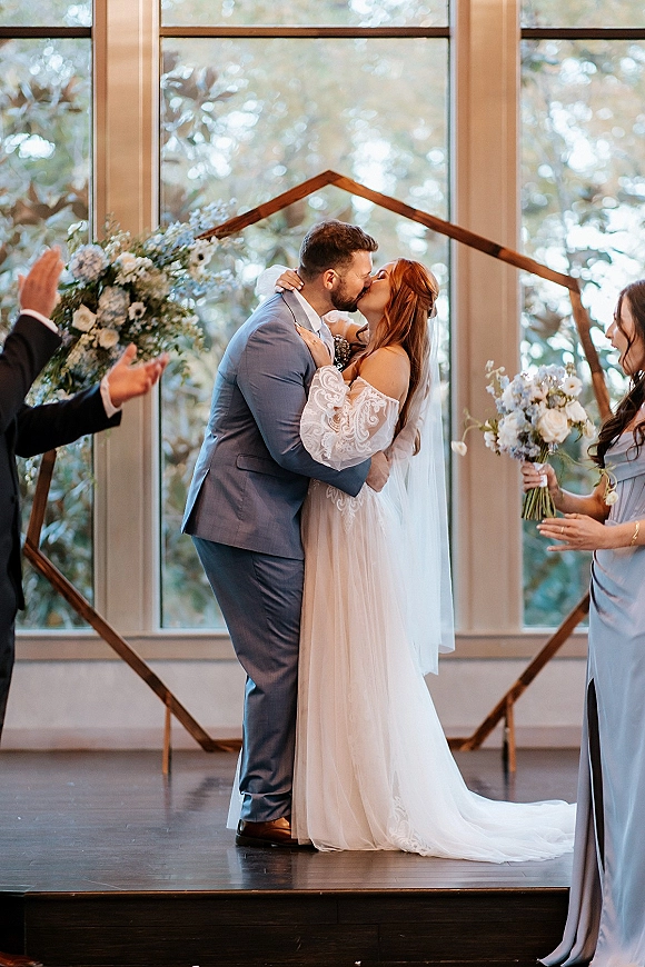Wedding kiss under a floral arch, bride in lace dress and veil embracing groom in a blue suit on an indoor stage by large windows.