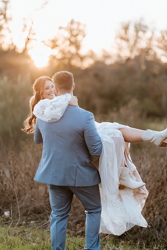 Couple portrait of groom carrying bride over his shoulder, her lace-sleeve wedding dress and bridal cowboy boots glowing in a sunset field