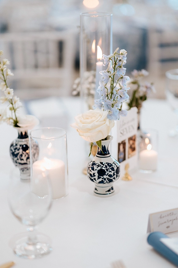 Reception tablescape with a wedding table centerpiece of white roses and blue delphinium in bud vases, pillar candles, and menu cards on white linens