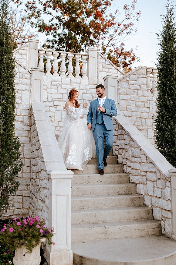 Couple portrait of bride and groom stairs, holding hands on a stone staircase as the bride in a lace dress carries champagne flutes