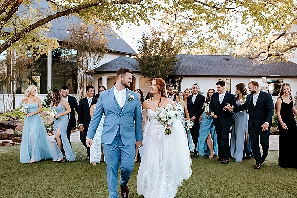 Wedding party portrait of the bride and groom walking hand in hand, bouquet and veil flowing, with bridesmaids and groomsmen on a garden lawn