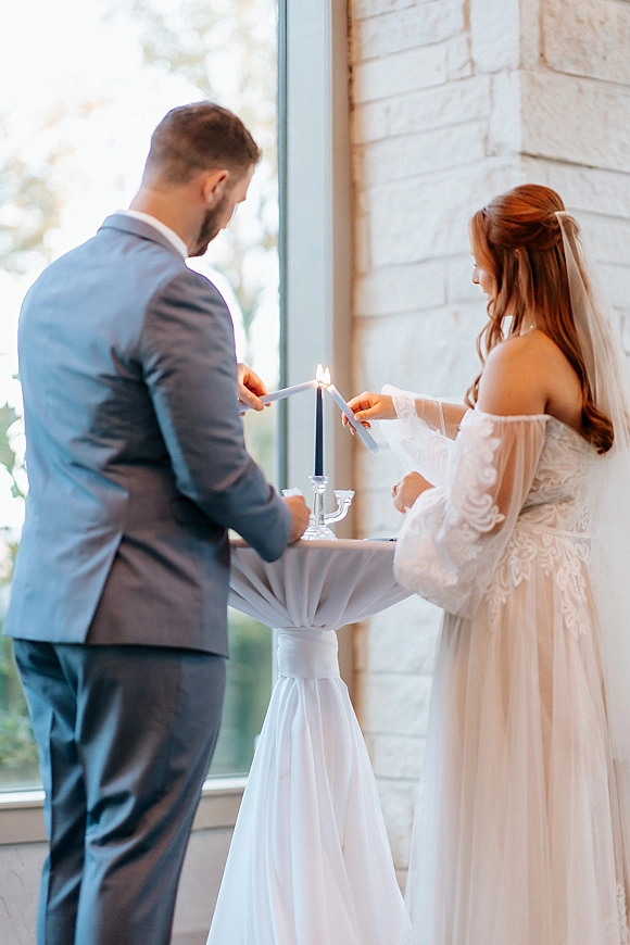Unity candle ceremony as bride in lace gown and groom in suit light tall tapers on a white table by a large window and stone wall
