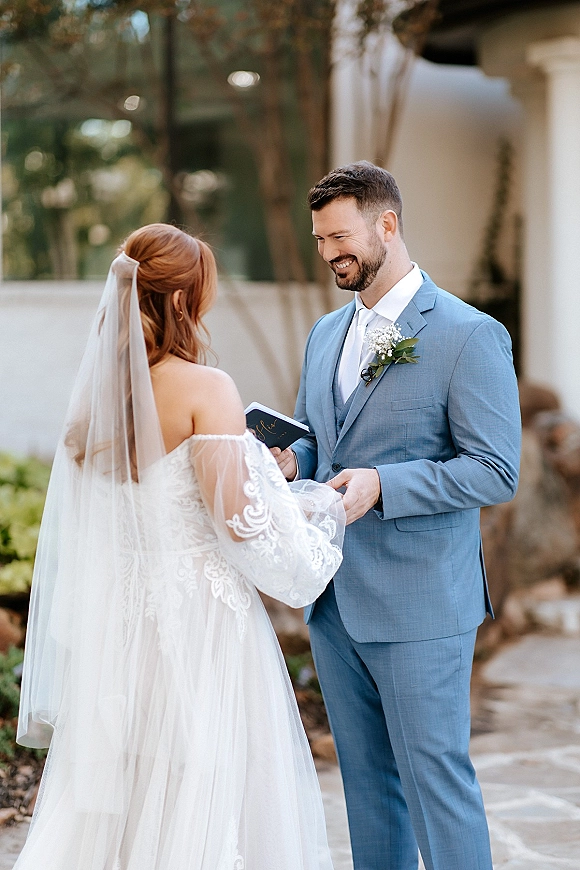 Wedding vows as the couple exchanging vows, holding hands while the groom reads from a vow book in an outdoor courtyard with trees