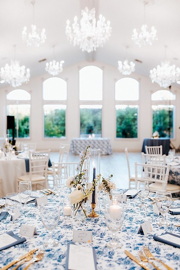 Reception tablescape with blue and white wedding table linens, white florals, hurricane vase, gold candlesticks, and candlelight under chandeliers