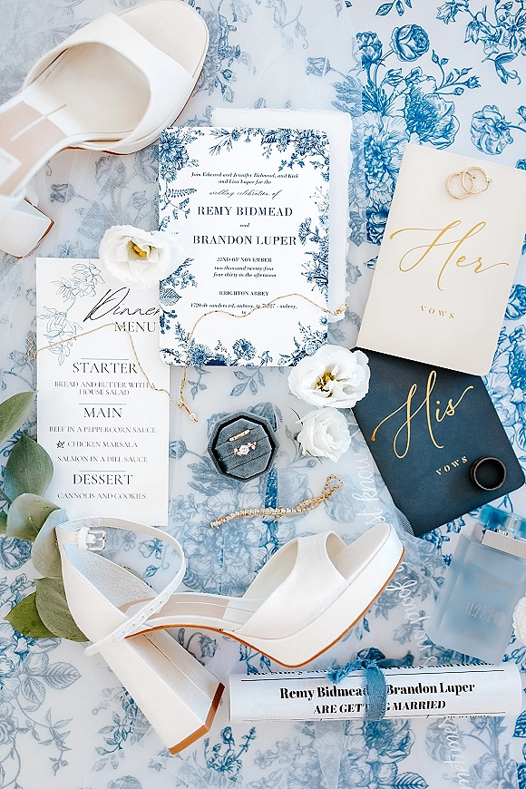 Wedding invitation suite with blue floral wedding invitations, menu and vow books beside rings, perfume and heels on lace and chinoiserie fabric backdrop