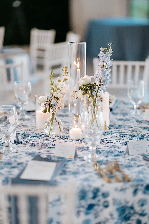 Reception tablescape with a blue and white wedding tablescape on toile tablecloth, bud vases of pale blooms and candlelight in a blurred room