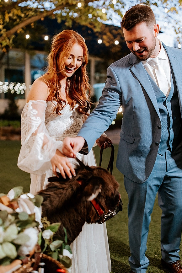 Couple portrait of bride and groom with dog, holding a leash as string lights glow over an evening lawn with trees behind